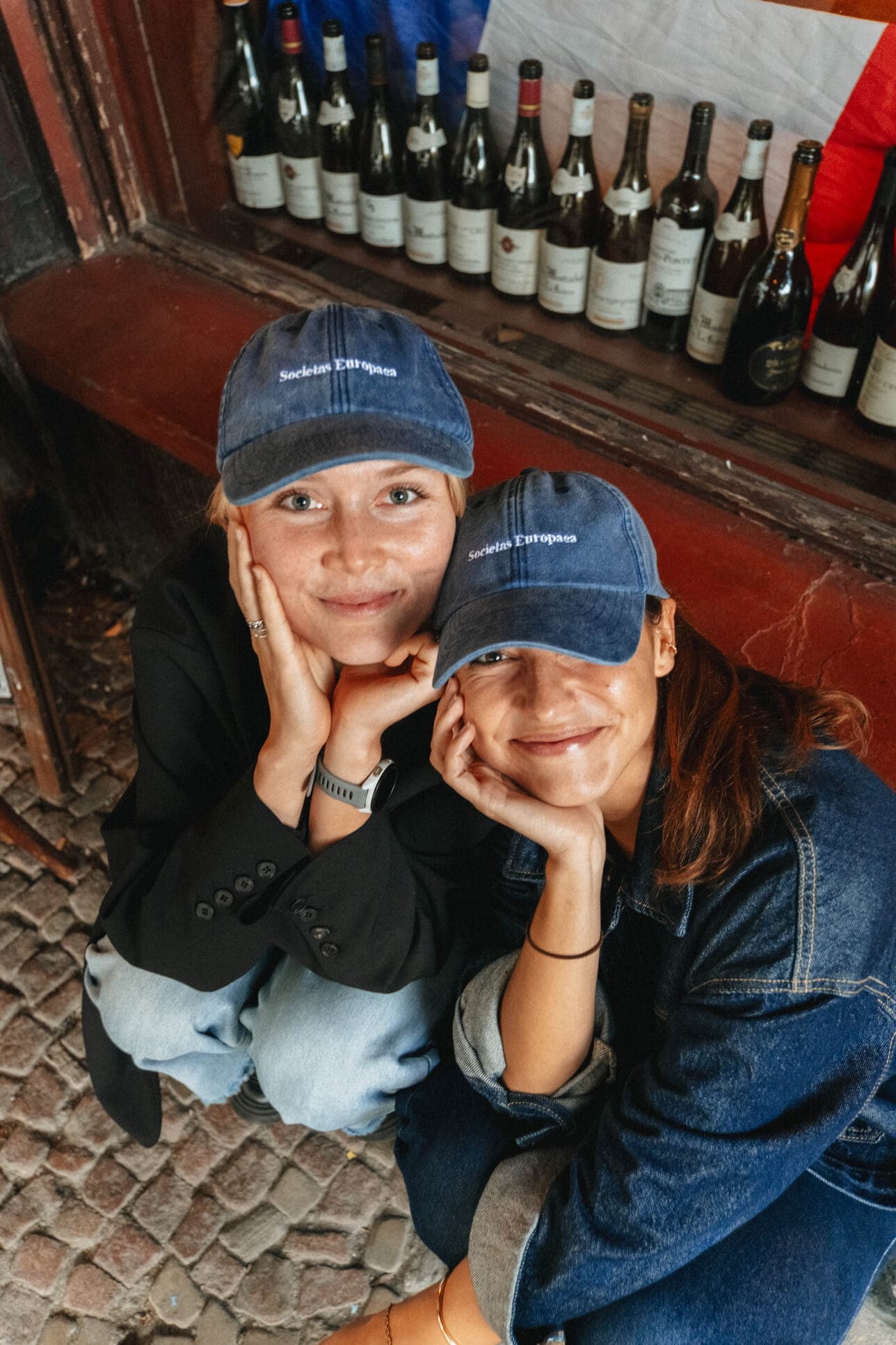 Two teammates in blue caps smiling beside a wine shop window.